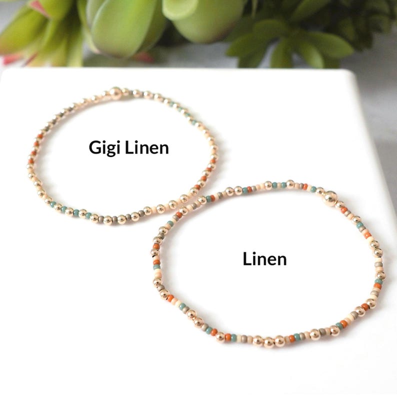 May include: Two beaded bracelets. One bracelet is gold with the text "Gigi Linen". The other bracelet is gold with the text "Linen". The bracelets have a mix of gold, beige, teal, and orange beads.