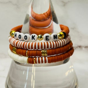 May include: Stack of orange and white beaded bracelets. One bracelet spells out "HOOK EM" with white letter beads. Gold and black accents are visible on the bracelets. The bracelets are stacked on a clear glass stand.