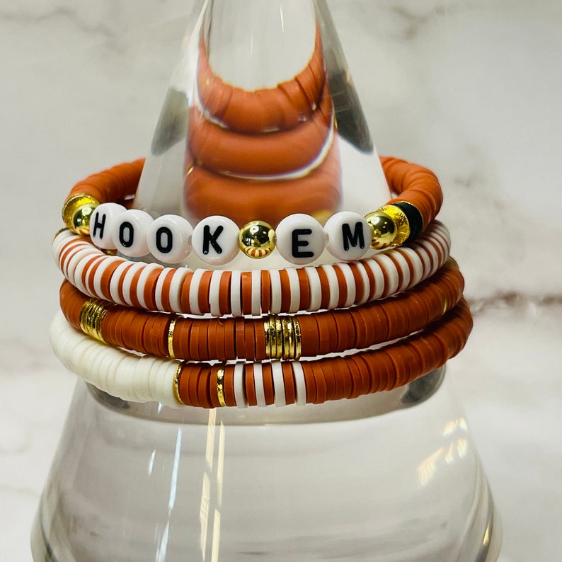 May include: Stack of orange and white beaded bracelets. One bracelet spells out "HOOK EM" with white letter beads. Gold and black accents are visible on the bracelets. The bracelets are stacked on a clear glass stand.