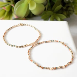 May include: Two beaded bracelets. One bracelet features gold-colored beads with accents of teal, orange, and cream-colored beads. The other bracelet is made of gold-colored beads. Both bracelets are on a white surface with green foliage in the background.