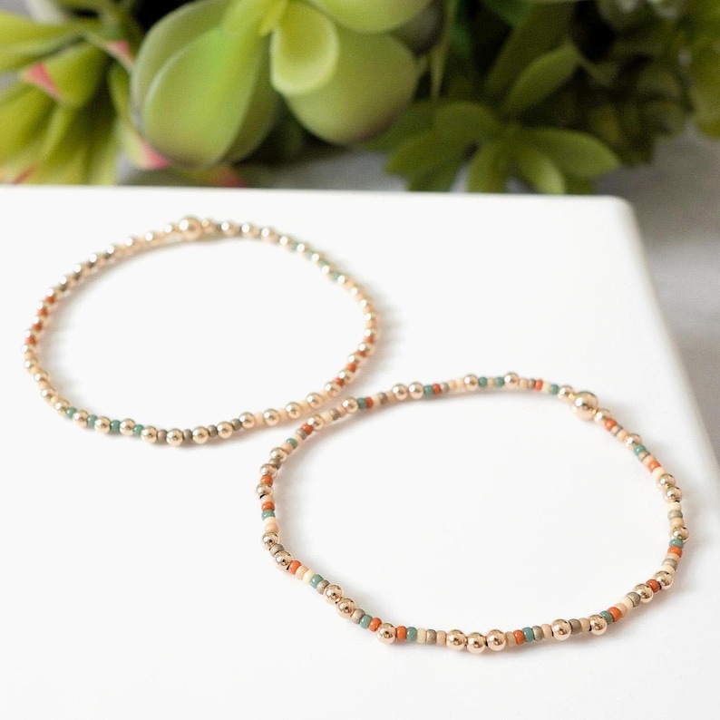 May include: Two beaded bracelets. One bracelet features gold-colored beads with accents of teal, orange, and cream-colored beads. The other bracelet is made of gold-colored beads. Both bracelets are on a white surface with green foliage in the background.