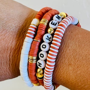 May include: Stack of five handmade bracelets. The bracelets feature a mix of terracotta, white, and gold accents. One bracelet spells out the word "HOOKEM" with white letter beads. The bracelets are made of flat, disc-shaped beads.