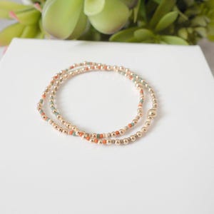 May include: Two gold beaded bracelets with small orange and teal accent beads. The bracelets are on a white surface with green foliage in the background. The bracelets are made of small gold beads.