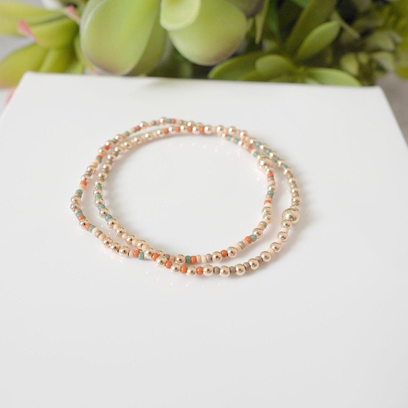 May include: Two gold beaded bracelets with small orange and teal accent beads. The bracelets are on a white surface with green foliage in the background. The bracelets are made of small gold beads.