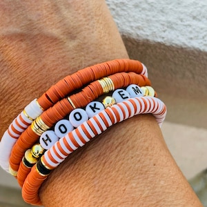 May include: A stack of beaded bracelets in shades of orange, white, and gold. One bracelet spells out the word "HOOKEM" with white letter beads. The bracelets are made of flat, disc-shaped beads and gold accents.