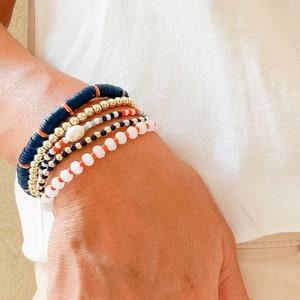 Puede incluir: Un conjunto de pulseras con cuentas negras, blancas, naranjas y doradas. Las pulseras están en la muñeca de una persona.