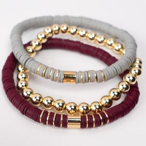 May include: Three stacked bracelets. The top bracelet is grey with gold beads and accents. The middle bracelet is a deep burgundy colour with gold beads and accents. The bottom bracelet is also burgundy with gold beads and accents.
