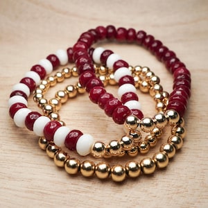 May include: Three bracelets stacked on top of each other. The top bracelet is red and white, the middle bracelet is gold, and the bottom bracelet is red and white.