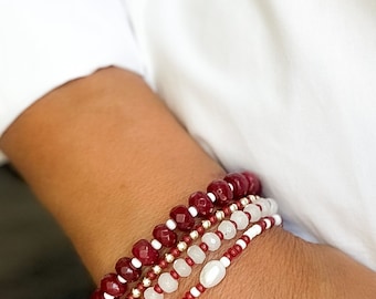 Könnte beinhalten: Ein Set aus vier Armbändern mit roten und weißen Perlen. Die Armbänder sind auf dem Handgelenk einer Person übereinander gelegt. Das oberste Armband besteht aus größeren, roten, facettierten Perlen mit weißen Akzenten. Das zweite Armband besteht aus kleineren, weißen Perlen mit roten Akzenten. Das dritte Armband besteht aus kleineren, weißen Perlen. Das unterste Armband besteht aus goldenen Perlen.