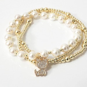 May include: Three gold beaded bracelets with white faux pearls. A small gold cat charm with clear crystals hangs from one bracelet. The bracelets are stacked on a white surface.
