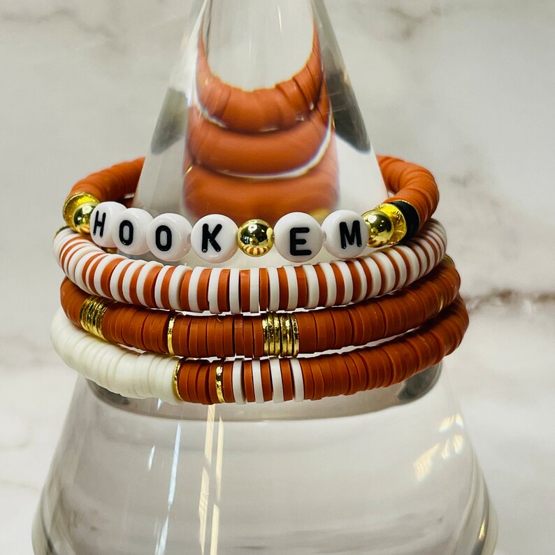 May include: Stack of bracelets in shades of orange, white, and gold. One bracelet spells out "HOOK EM" with white letter beads. The other bracelets have a mix of solid and striped designs. The bracelets are made of flat, disc-shaped beads.