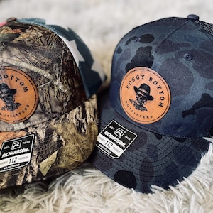 May include: Two camouflage trucker hats with a black mesh back. The hats have a brown leather patch with the text "Soggy Bottom Outfitters" and a skull graphic. The hats are made by Richardson.