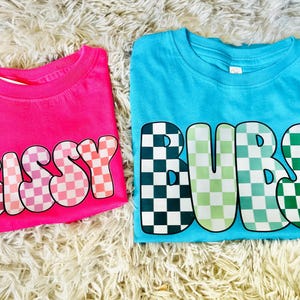 May include: Two t-shirts with checkered print designs. The pink shirt says "Sissy" and the blue shirt says "Bubs".