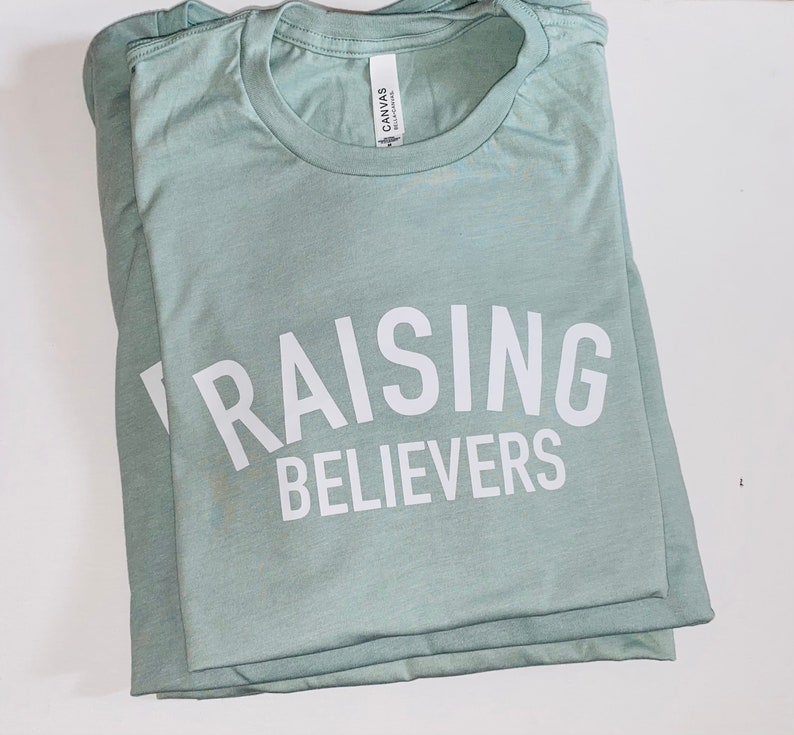 May include: A light green t-shirt with the text "RAISING BELIEVERS" printed in white.