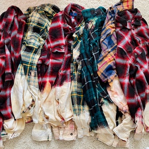 May include: A collection of plaid flannel shirts in various colors, including red, black, green, and yellow. Each shirt has a bleached, distressed bottom half, creating a unique, worn look. The shirts are button-down with long sleeves.