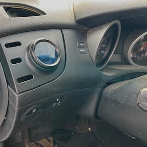 May include: A black automotive dashboard interior featuring a circular gauge mounted in a custom vent pod. The dashboard includes horizontal air vent slats, a trip reset button, and a portion of the instrument cluster with a speedometer visible.