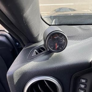 May include: A black gauge mounted on a car's dashboard, displaying a red needle against a black background. The dashboard is textured black plastic, with air vents and other controls visible. The gauge is cylindrical and appears to be aftermarket.