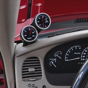 May include: A dual gauge pod mounted on a vehicle dashboard featuring two circular gauges with silver bezels. The top gauge displays oil pressure with a red needle, and the bottom gauge displays boost pressure with a red needle and black face.