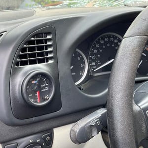 May include: Close-up of a car's dashboard with a black and white speedometer, a black steering wheel, and a small black gauge with a red needle. The gauge reads "PSI" and "1000" and has a white background with black text.
