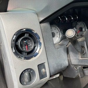 May include: Close-up of a car's dashboard with a gauge reading 25 PSI. The gauge is mounted in a silver bezel with a black background. The dashboard is gray and has a variety of controls.