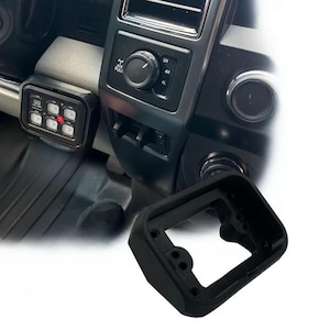 May include: Black rectangular automotive switch panel with six illuminated buttons, mounted on a vehicle's dashboard. A black mounting bracket is in the foreground. The dashboard features control knobs and vents.