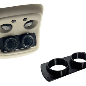 May include: A beige car interior component with two circular vents and control knobs. A separate black plastic piece with two circular openings sits next to it. Automotive parts.