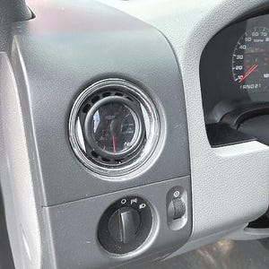 May include: Close-up of a gray dashboard interior with a round gauge and a black control knob. The gauge has a chrome bezel and a red needle. The control knob has symbols for lights.