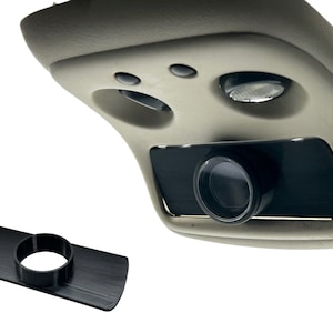 May include: A black plastic adapter for a car camera. The adapter is designed to fit into the overhead light fixture of a car. The adapter has a round hole in the center.
