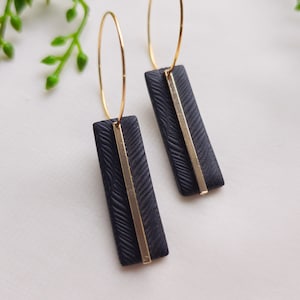 May include: A pair of black hoop earrings with gold accents. The earrings feature a rectangular shape with a textured surface and a thin gold line running down the center.