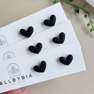 May include: Close-up of black heart-shaped stud earrings on white cards. The cards have a logo and the text "ALLBYBIA EST. 2022". The earrings are arranged on the cards, with a few visible. Green leaves are in the background.