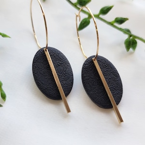 May include: A pair of gold hoop earrings with black oval-shaped charms. The charms have a textured pattern and a gold bar extending from the bottom.