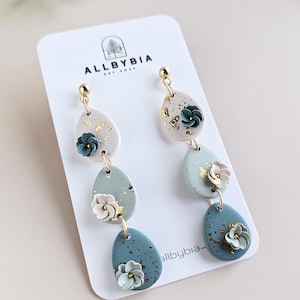 Puede incluir: Pendientes colgantes hechos a mano con temática floral. Cada pendiente presenta tres segmentos en forma de lágrima en tonos azules y blancos, adornados con pequeños detalles florales y detalles de pan de oro. Los pendientes se exhiben en una tarjeta blanca con la marca "ALLBYBIA".