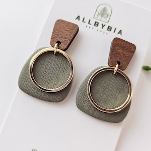 May include: A pair of earrings featuring a green leather and wood design. The earrings have a gold circle and a brown wood piece. The earrings are displayed on a white card with the text "ALLBYBIA EST. 2022" and "@allbybia_" printed on it.