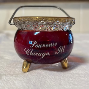 May include: A small red glass cauldron with a gold rim and a silver handle. It has three gold feet and says "Souvenir Chicago, Ill." on the side.