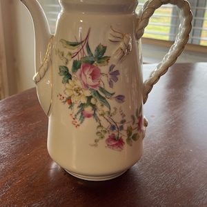 May include: A white porcelain teapot with a gold trim and a floral design. The teapot has a rope-like handle and a spout.