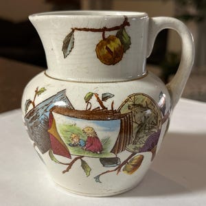 May include: A white ceramic pitcher with a handle, decorated with a colorful floral pattern and a scene of two children playing in a field. The pitcher has a gold trim around the top.
