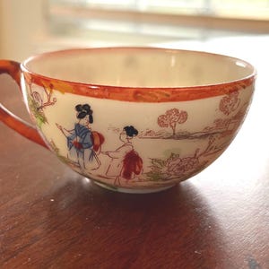May include: A white teacup with a red rim and a floral design featuring two women in traditional Japanese clothing. The cup has a handle and is sitting on a wooden surface.
