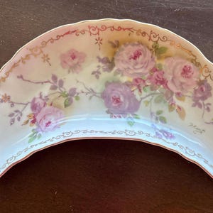 May include: A crescent-shaped, antique porcelain dish with a floral design. The dish features a delicate pattern of pink and purple roses with green leaves, accented by gold trim along the edges. The background is a soft, off-white color.