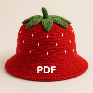Può includere: Un cappello rosso all'uncinetto a forma di fragola, con uno stelo e foglie verdi. Il cappello presenta dettagli di semi bianchi e le lettere bianche "PDF".