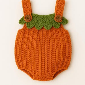 May include: Hand-crocheted orange baby romper with green scalloped trim and brown buttons. The romper has a pumpkin design, ideal for a baby's Halloween costume or autumn-themed photoshoot. The straps are attached with two brown buttons.