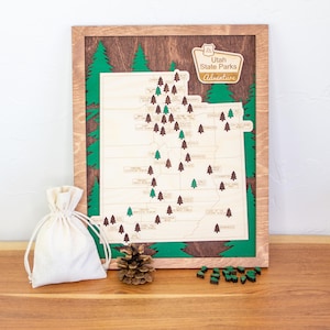 May include: Wooden map of Utah State Parks with a brown frame and green accents. The map features small wooden tree markers and the text "Utah State Parks Adventure". A small canvas bag, pine cone, and tree-shaped game pieces are in front of the map.