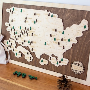 May include: Wooden map of the United States with state names and green and brown tree markers. The map is mounted on a dark brown wooden background and framed in light wood. The words "National Park Adventures" are on a small wooden sign.