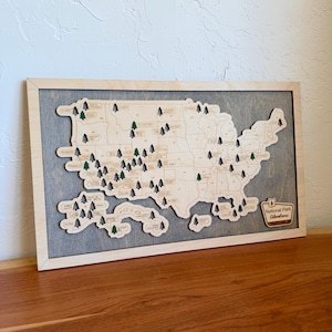 May include: A framed wooden map of the United States featuring laser-cut state borders and small tree icons marking national park locations. A badge in the bottom right corner displays the text National Park Adventures in script.
