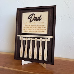 May include: Wooden baseball-themed sign with the word "Dad" in a cursive font. The sign includes a heartfelt message and personalized baseball bat cutouts with names. The frame is dark brown, and the bats are light brown.