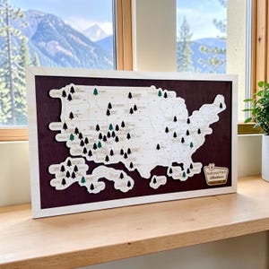 May include: A rectangular wooden wall art piece featuring a light-colored map of the United States mounted on a dark purple background. The map includes small black and green tree icons marking national park locations and a sign reading National Park Adventures.