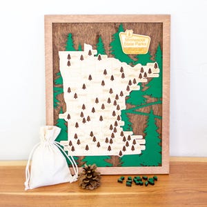May include: Wooden map of Minnesota State Parks with a green and brown design. The map features small wooden tree markers and a white drawstring bag. A pine cone and small green wooden pieces are also present.
