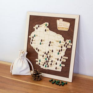 May include: A wooden map of Wisconsin State Parks featuring a dark brown background and a light wood cutout of the state. The map includes small tree-shaped markers, a pinecone, a small fabric drawstring bag, and a sign labeled Wisconsin State Parks Adventure.