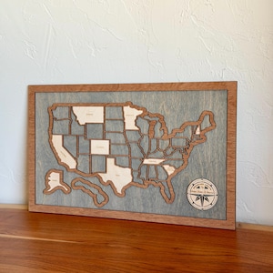 May include: A rectangular wooden wall art piece featuring a laser-cut map of the United States of America. The map is mounted on a grey-stained wooden board with a natural wood frame. A circular compass rose emblem is in the bottom right corner.