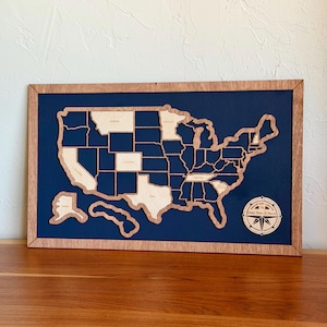 May include: A rectangular wooden wall map of the United States featuring light-colored wooden state cutouts mounted on a dark blue background within a wooden frame. A circular compass rose emblem in the bottom right corner reads Adventure Awaits.