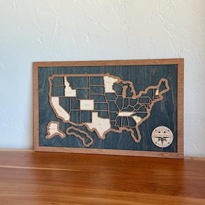 May include: A rectangular wooden wall art piece featuring a map of the United States with light-colored wood state cutouts set against a dark blue-stained background. A circular compass rose emblem in the bottom right corner reads Adventure Awaits.